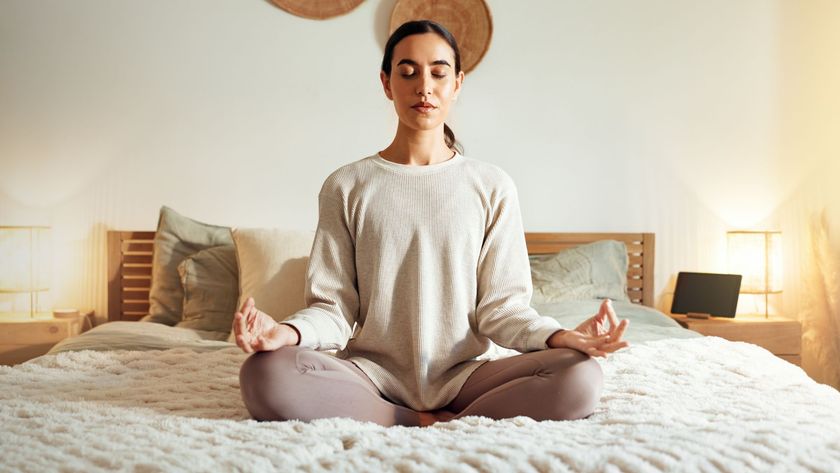 A woman sits cross-legged on a bed with her eyes closed and her fingers held in a meditative position.
