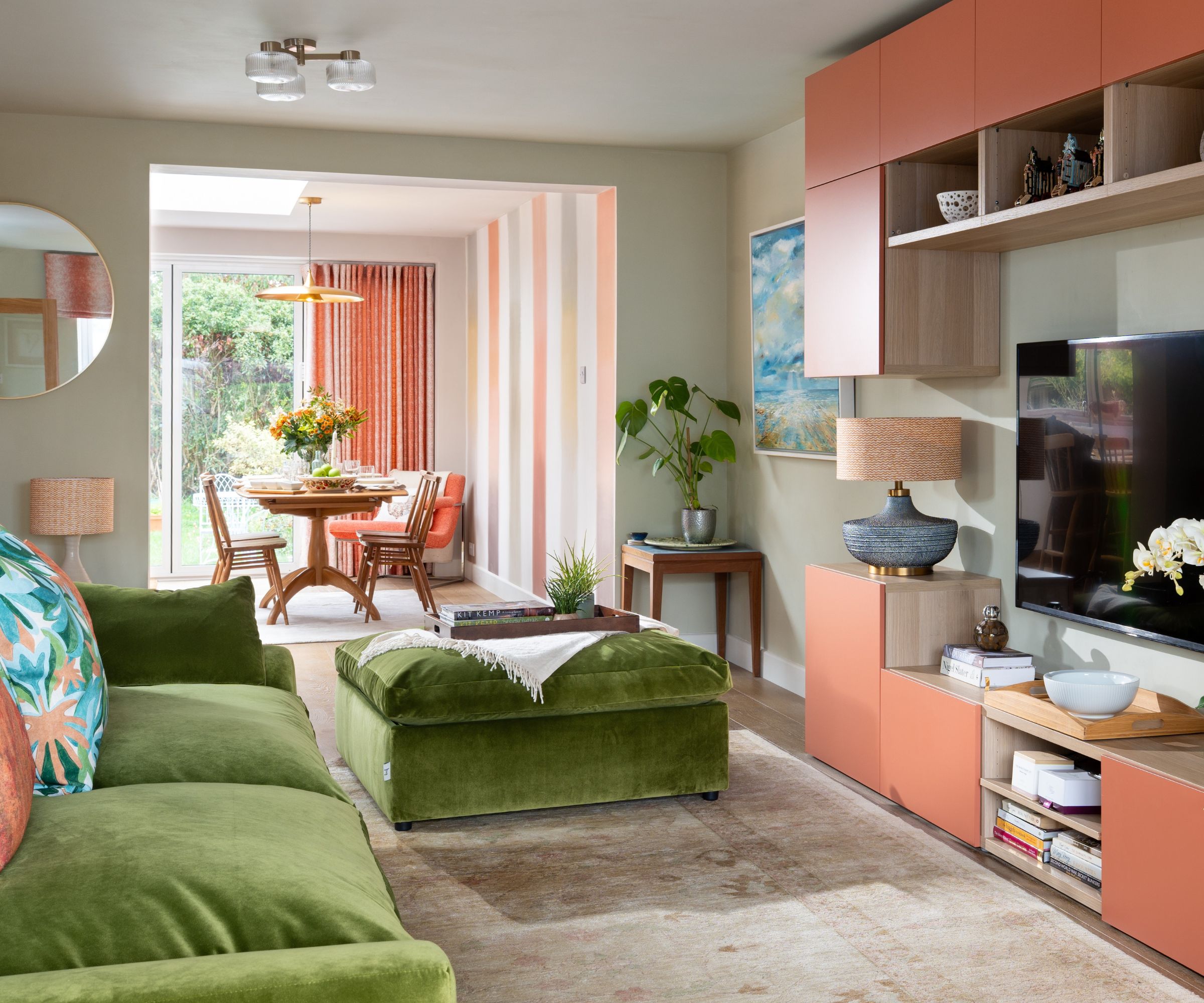 Living room with built-in storage cubby boxes painted in Naperon by Farrow &amp;amp; Ball, a green velvet sofa and matching ottoman, a beige rug, a large TV, with an open plan dining room in the background.