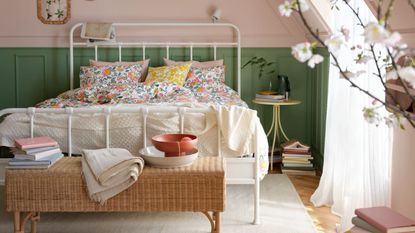 A attic bedroom space with pink and green panelled walls, a large white metal framed double bed with colorful, floral bedding, and a rattan ottoman at the end of it with a stack of books, white blanket, and two stacked bowls in white and orange. Beneath the bed is a white rug and wooden flooring, and to the right is a circular bedside table, stacks of books, large window with sheer white curtains, and a blurred cherry blossom plant in the foreground.