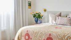 A white bedroom with a patterned duvet, a group of pillows, a gold wall sconce, a vase with peach flowers, a speckled mini vase, a framed painting, and a set of sheer curtains