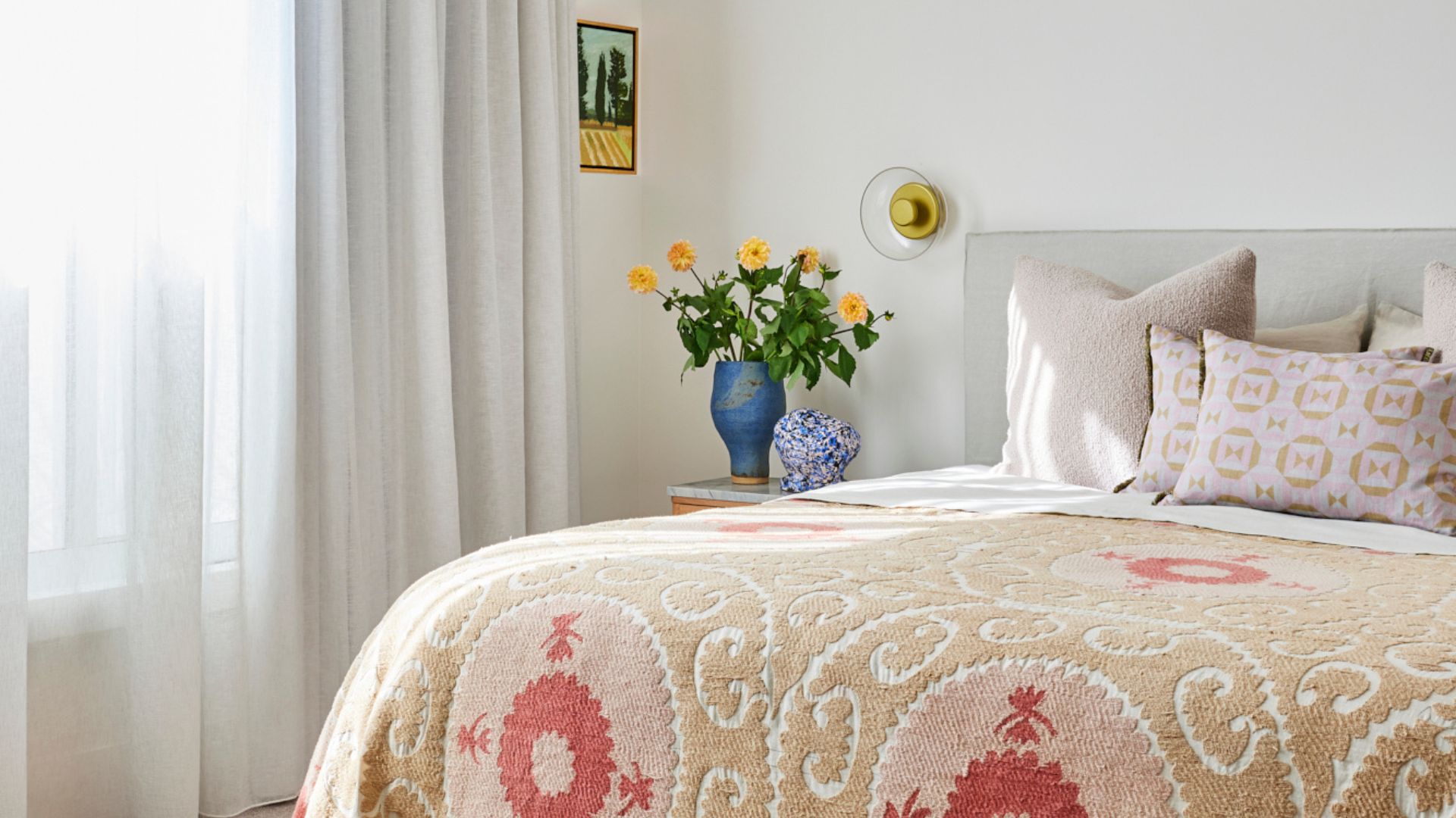 A white bedroom with a patterned duvet, a group of pillows, a gold wall sconce, a vase with peach flowers, a speckled mini vase, a framed painting, and a set of sheer curtains