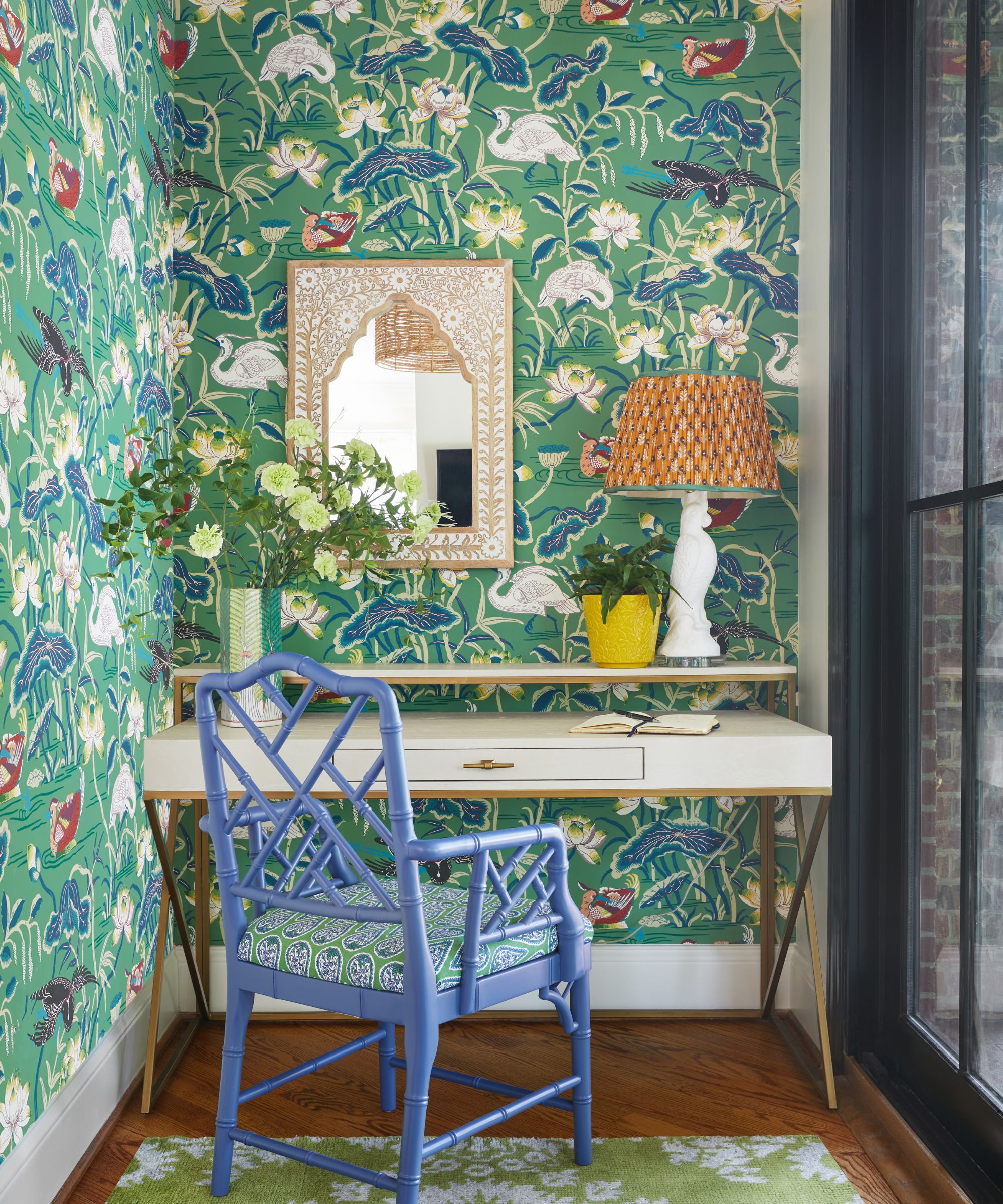 Home office with green floral wallpaper, a white desk, a blue painted cane chair, an inlay mirror on wall and a vase of green flowers