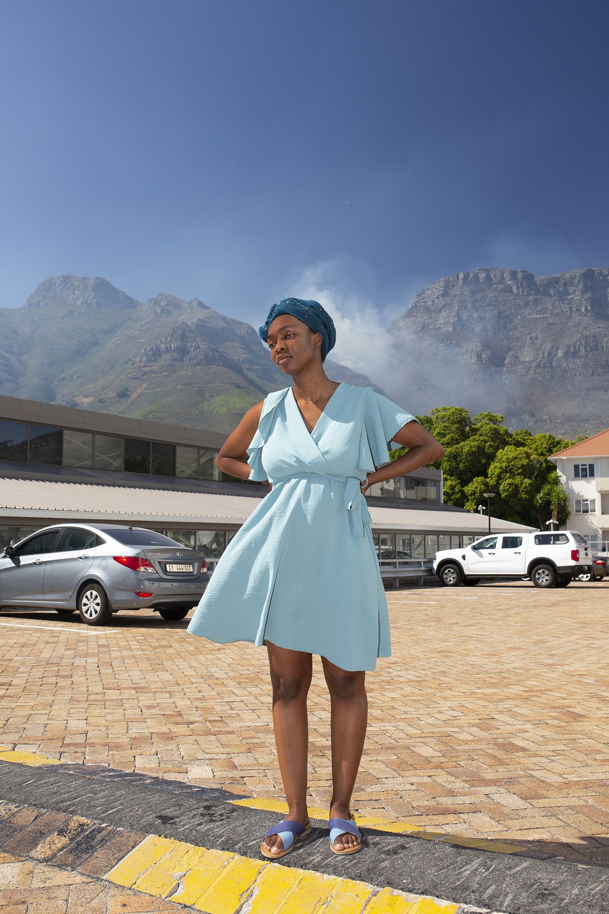 A woman in a light blue dress and headwrap stands confidently on a cobblestone street against a mountainous backdrop. Cars are parked nearby under a clear sky