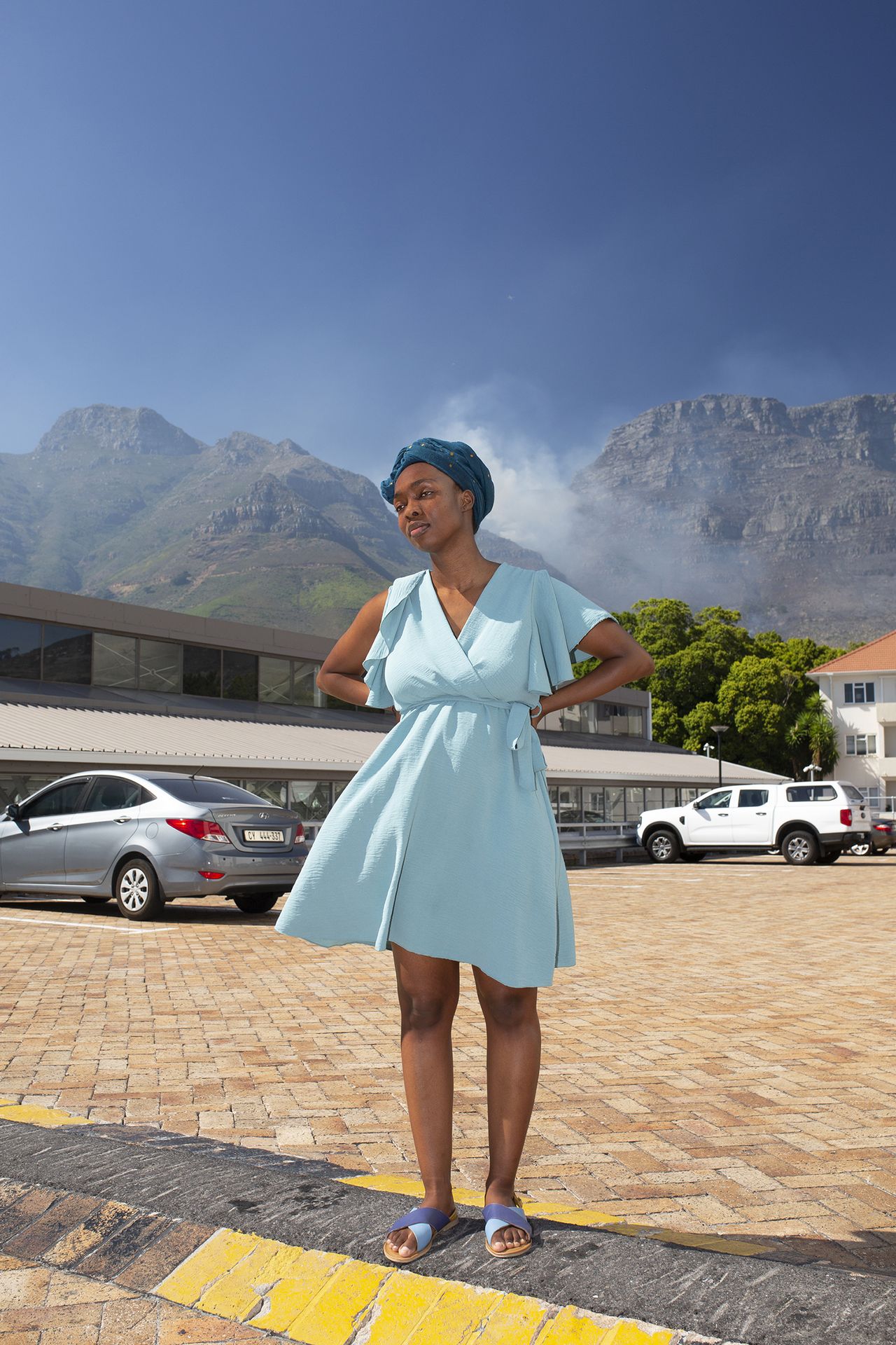 A woman in a light blue dress and headwrap stands confidently on a cobblestone street against a mountainous backdrop. Cars are parked nearby under a clear sky