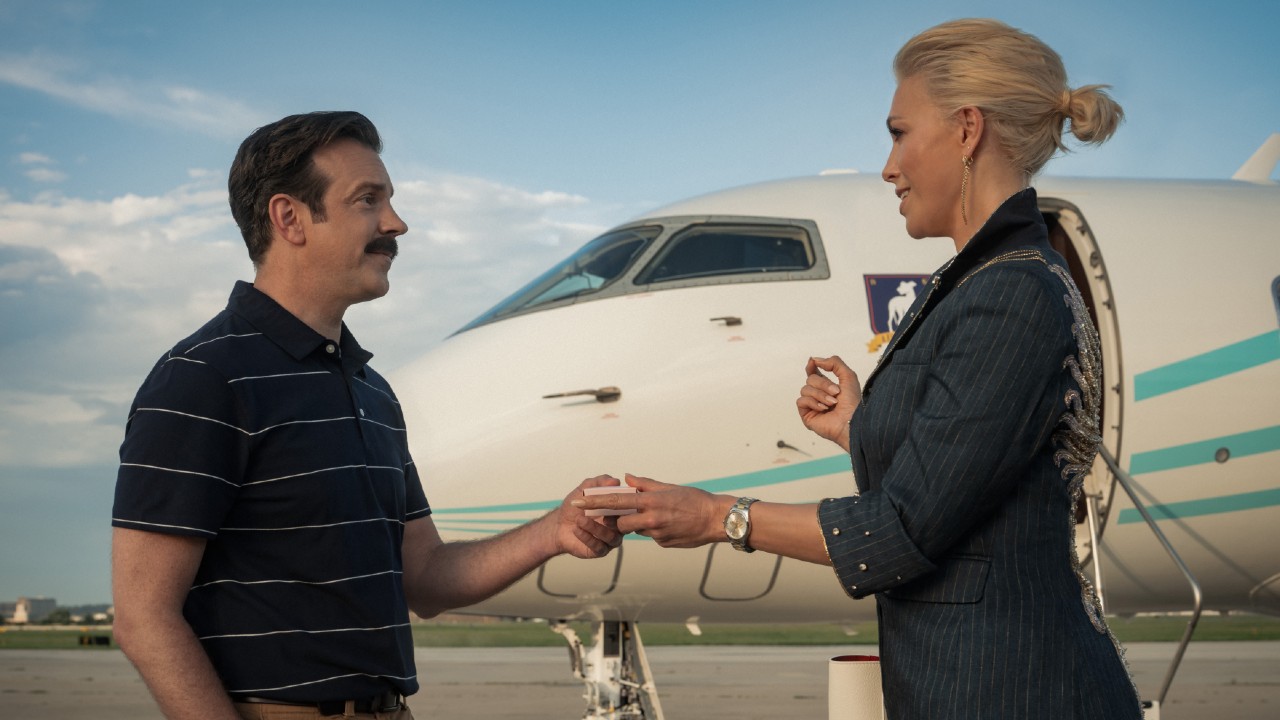 From left to right: Jason Sudeikis and Hannah Waddingham standing outside of a plane holding a box of biscuts. 