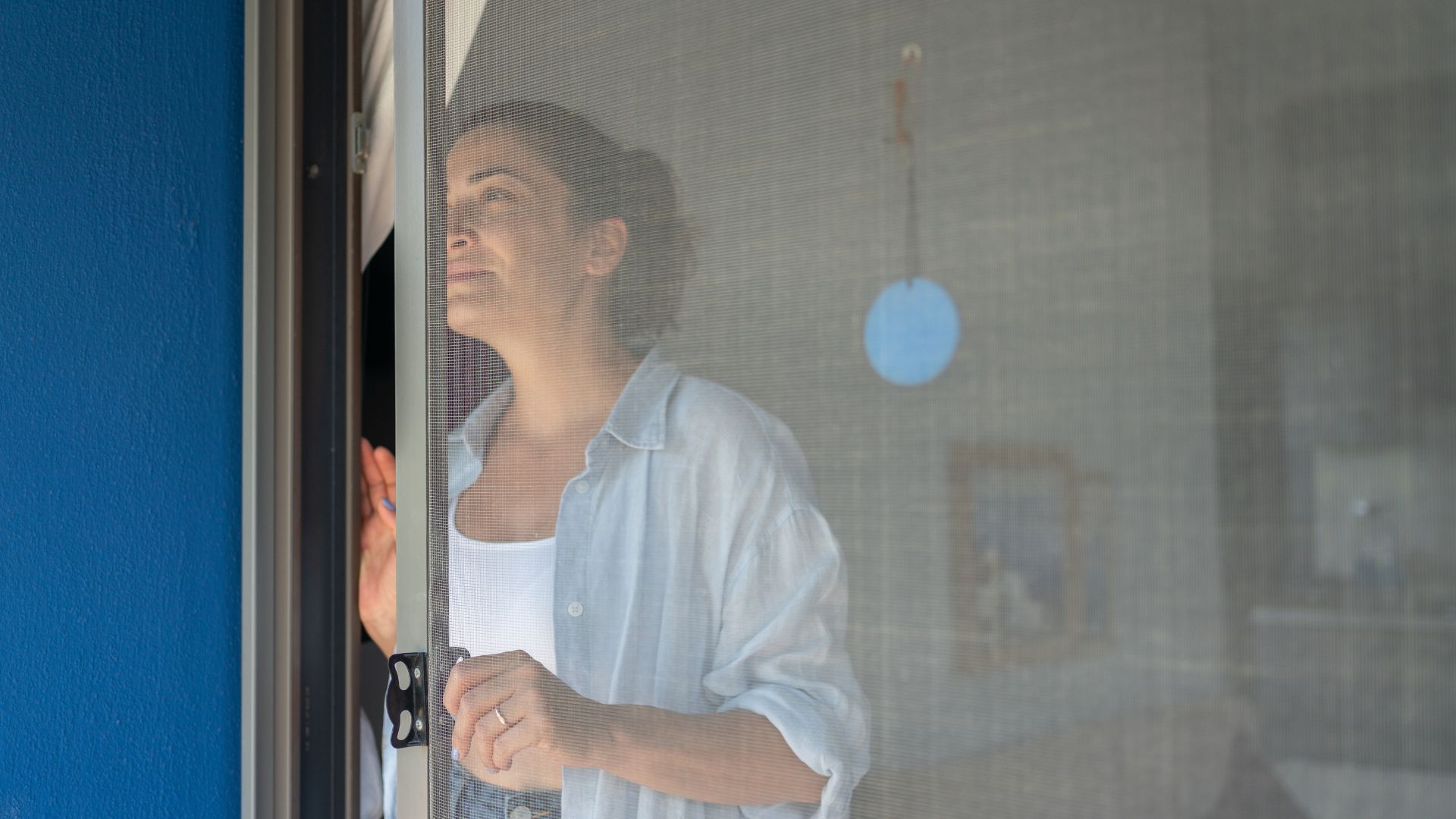 A brunette woman opens a window with a mosquito screen.