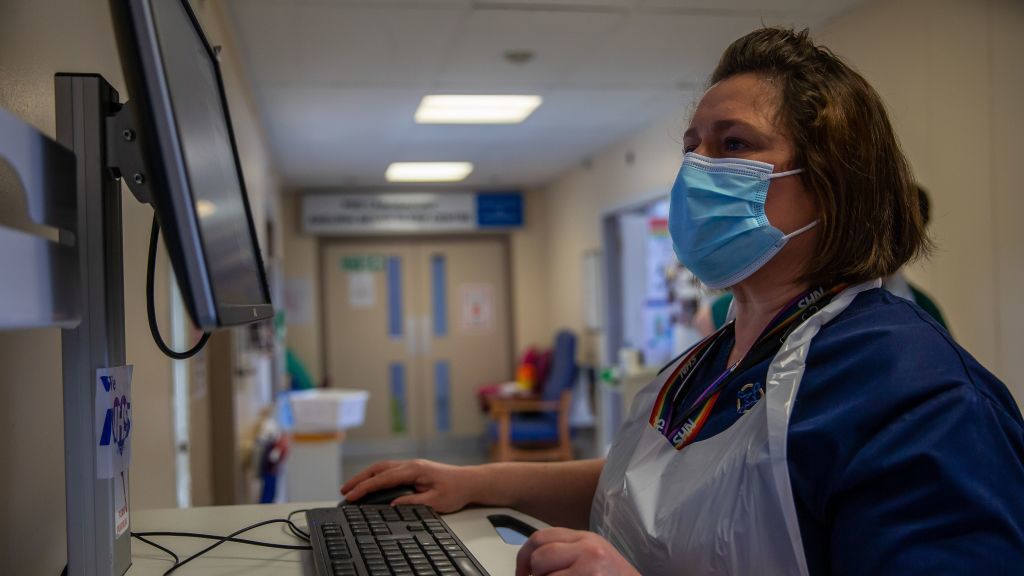 An NHS nurse wearing a face mask using a computer in a hospital corridor