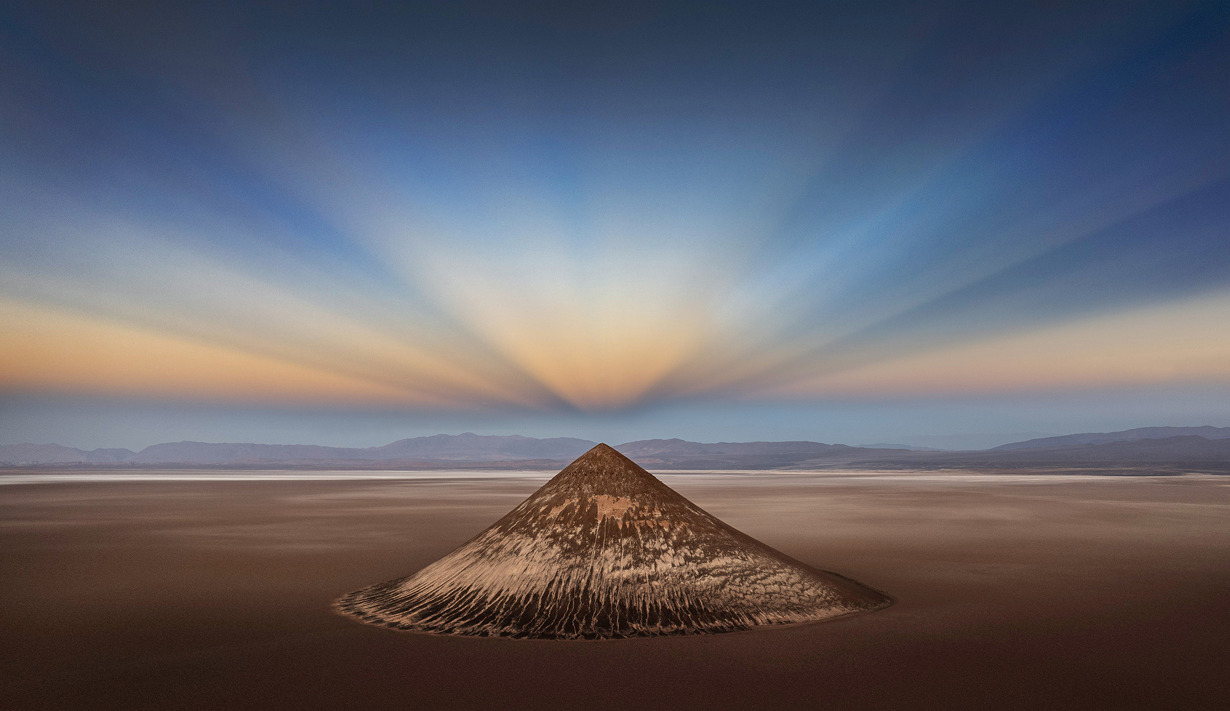 A solitary, conical volcanic mound rises from the barren landscape, bathed in soft, radiant light at dusk