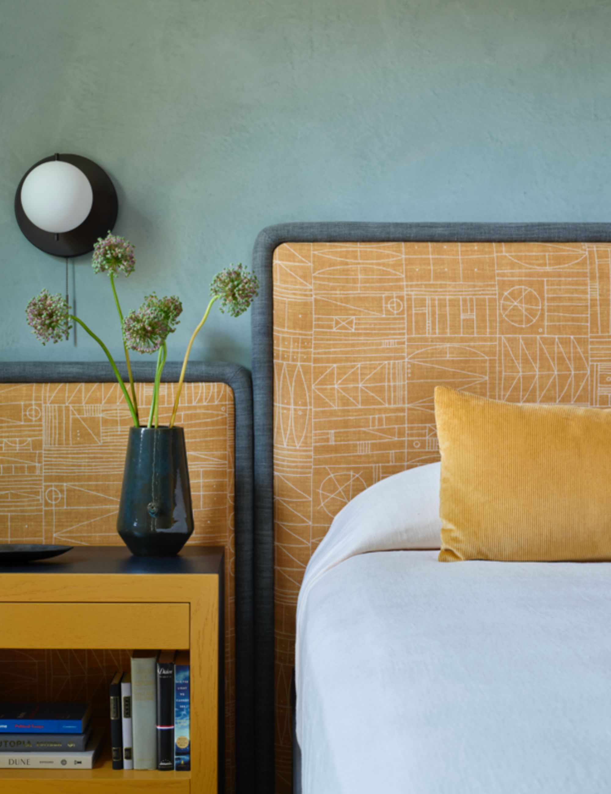 Teal colored bedroom with yellow printed headboard design and matching side table, complete with flowers, black plated side-light and white bed linen with a yellow cushion on top