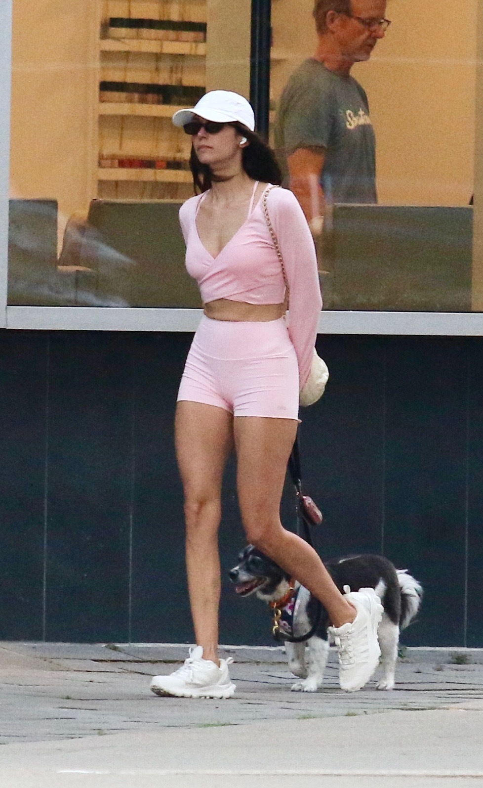 Nina dobrev wears a pink long-sleeve, pink alo shorts, a white baseball cap, and white sneakers.