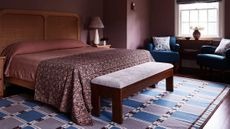 Burgundy bedroom with dark burgundy walls, rattan headboard, burgundy bedding, bench, purple and blue rug and window with two navy armchairs