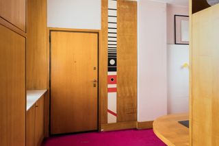 Wooden door, wooden cupboards, wooden table, pink carpet and abstract art on the wall.