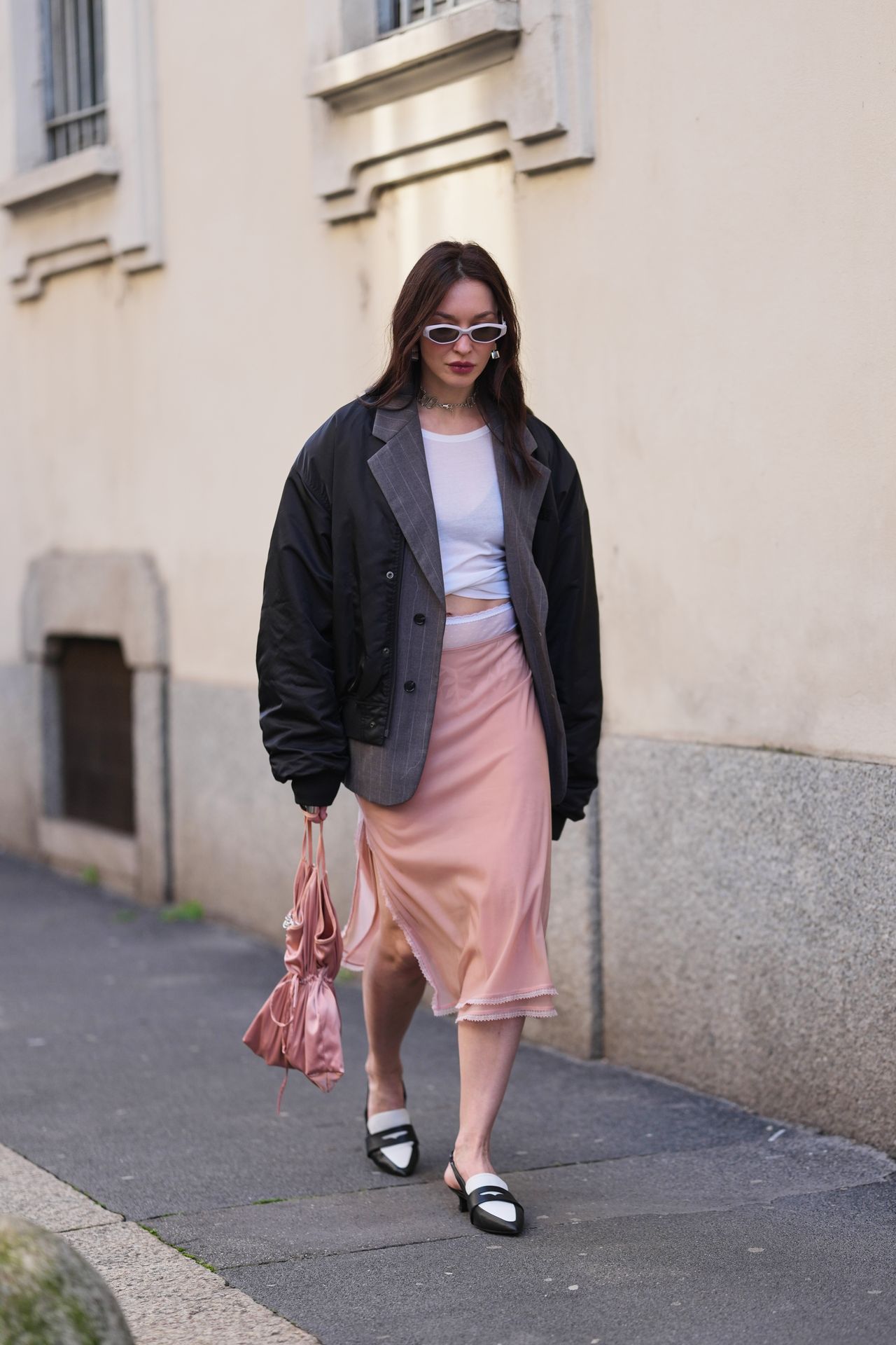MILAN, ITALY - FEBRUARY 26: A guest wears long dark brown hair worn loose with a center part, white silver cat-eye sunglasses, silver-tone pendant earrings, a silver chain necklace, a pink satin drawstring pouch bag, a black padded jacket, a gray pinstripe pattern blazer jacket, a white cropped wrap T-shirt, a blush pink satin slip midi skirt, black and white bicolored leather pointed-toe slingback pumps shoes, outside Anteprima, during Milan Fashion Week - Womenswear Fall/Winter 2026/2027, on February 26, 2026 in Milan, Italy (Photo by Edward Berthelot/Getty Images)