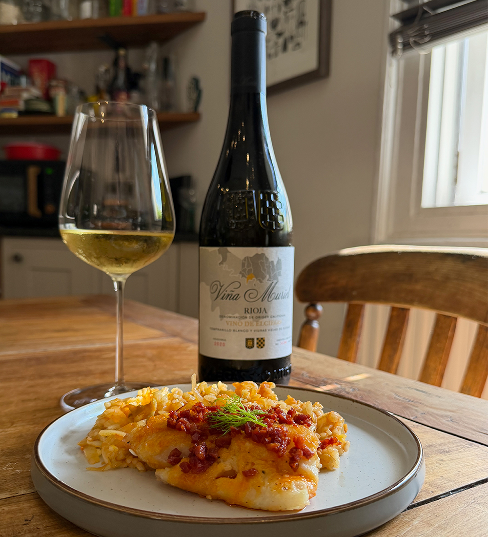Bottle of white wine with glass of white wine and plate of hake, rice and chorizo