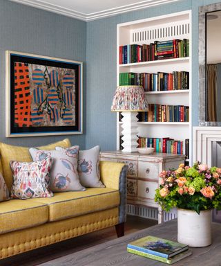 blue living room with yellow couch, patterned couch pillows, ornate sideboard, bookcase, modern artwork, flowers, coffee table