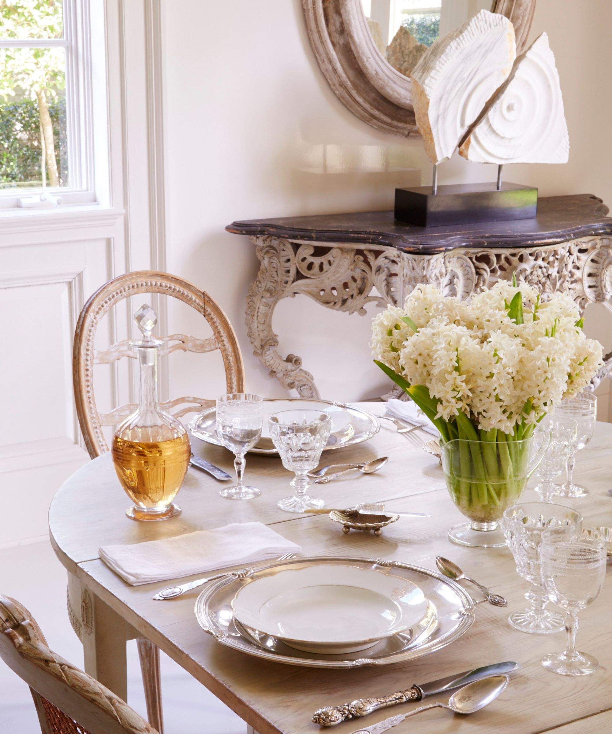 Dining room with traditional table setting including silver cutlery, china plates, and glassware. Console with decorative statue in the background