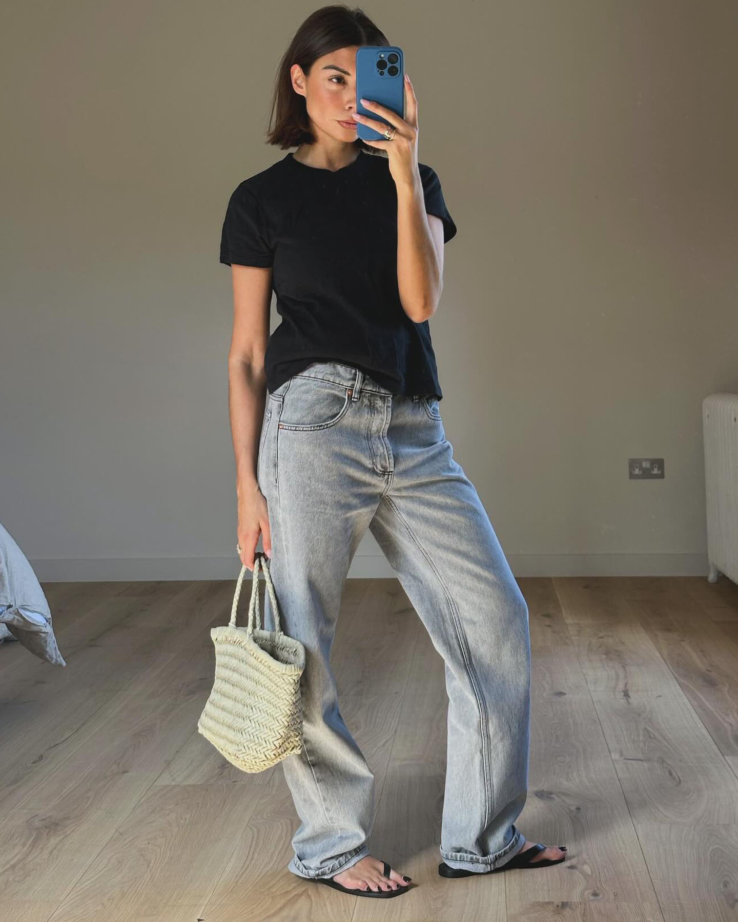 British influencer Marianne Smyth poses for a mirror selfie with a short bob haircut, a blue phone case, black T-shirt, baggy jeans, and neutral Dragon Diffusion tote bag.