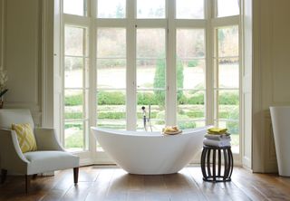 A white bath in a large window with an armchair and wooden stool with towels on it