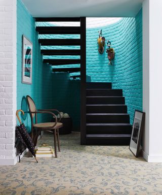 Characterful blue hallway, blue and cream patterned carpet, facing dark wood staircase, bright blue feature wall on stairwell, white painted wall and brick walls near stairway, decorated with paintings and ornaments on wall, traditional dark wood chair