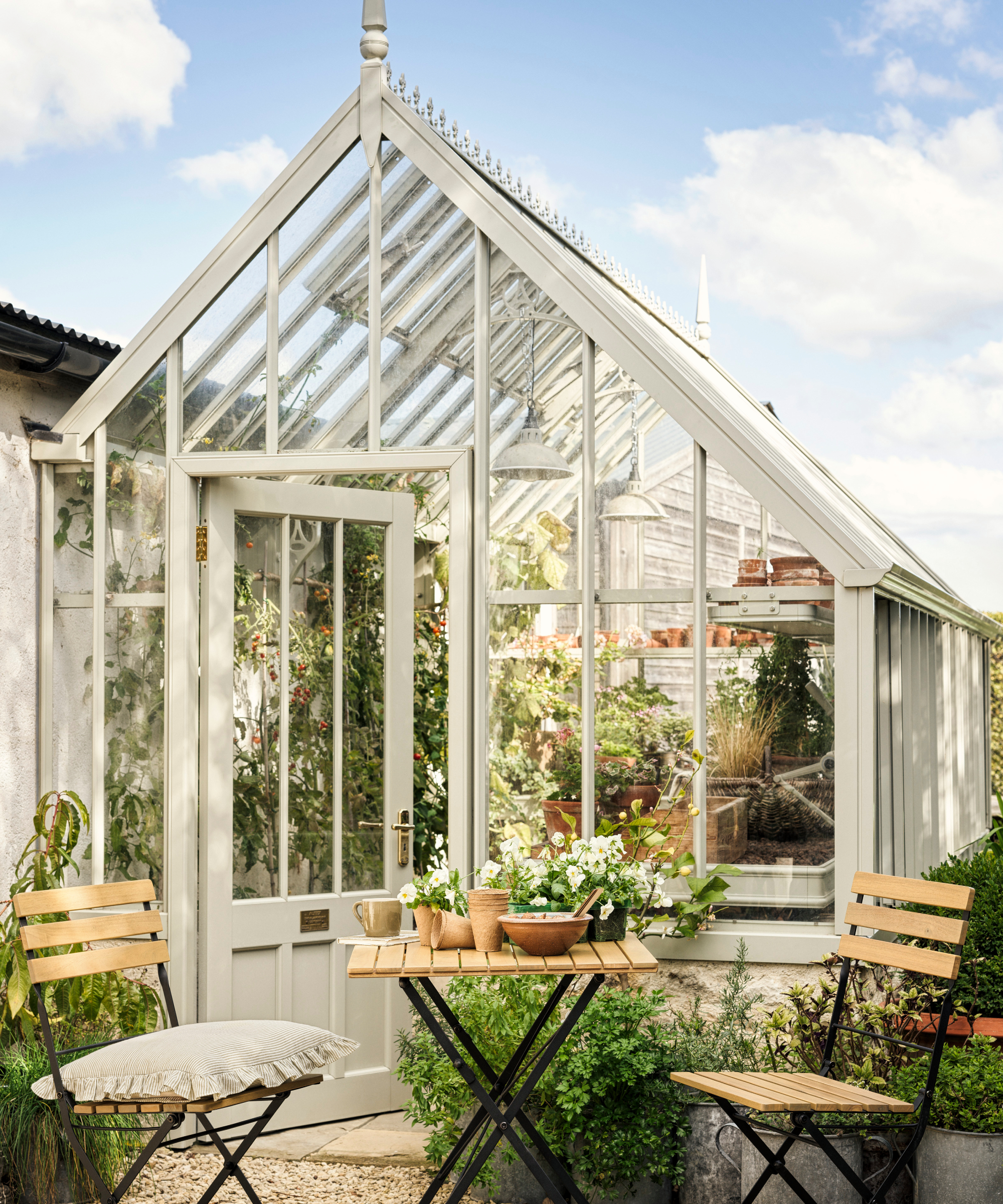 large greenhouse in cottage garden with pale green gothic like structure and wooden slatted bistro table and chairs on patio area in front of greenhouse