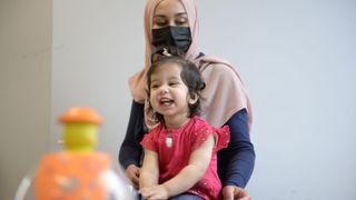 a smiling toddler wearing a red shirt sits in front of her mother who's wearing a black medical mask and pink head scarf
