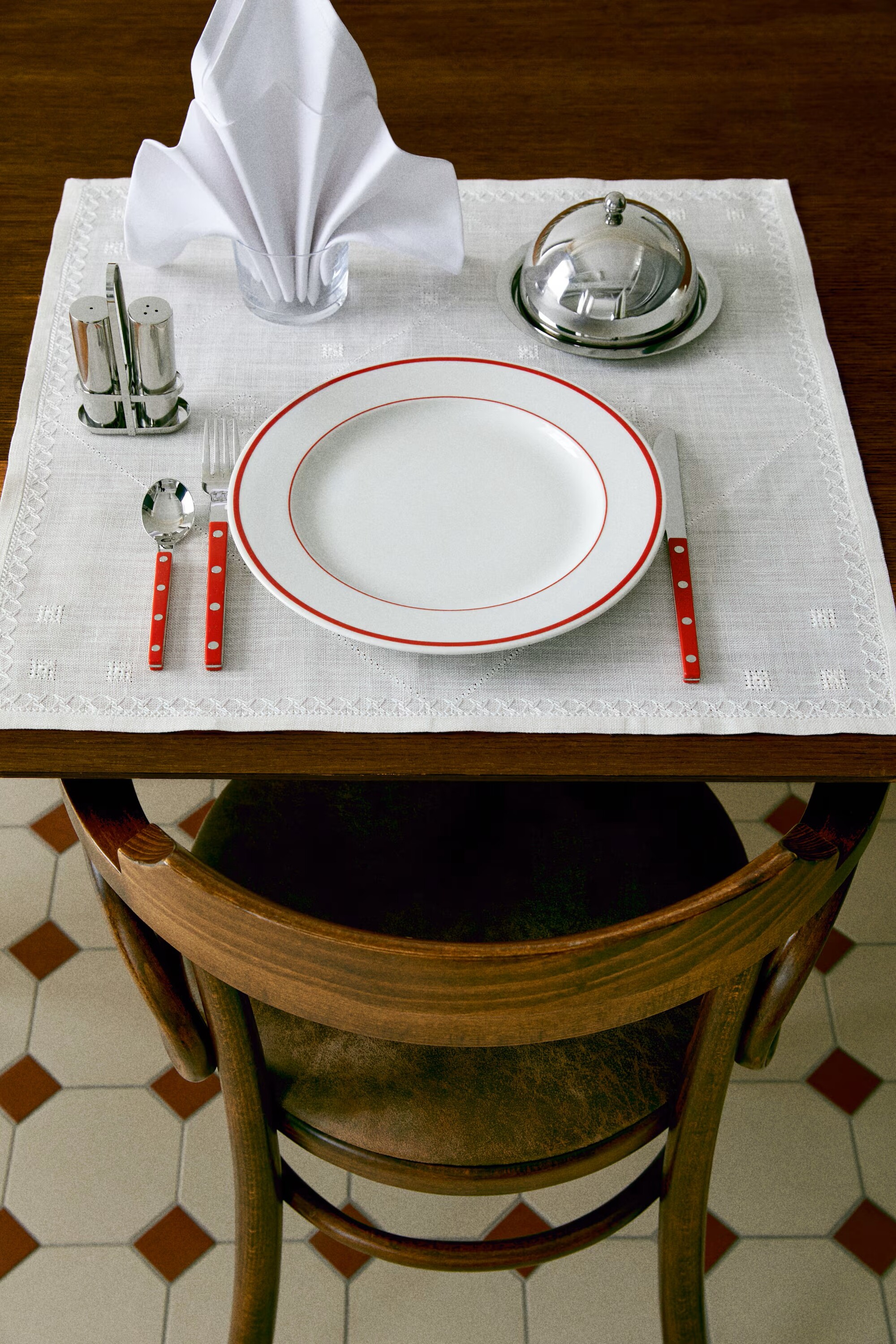 Image of a wooden table for two in a diner setting with a white tablecloth, a white plate with red trim, red utensils, and a silver butter dish