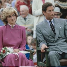 Prince Charles wearing a gray suit and Princess Diana in a pink suit holding flowers sitting in chairs next to each other with a crowd behind them