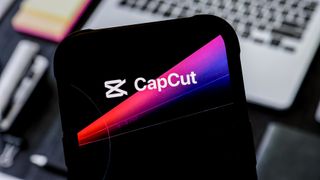 CapCut logo on a smartphone being held above a desk with laptop in the background