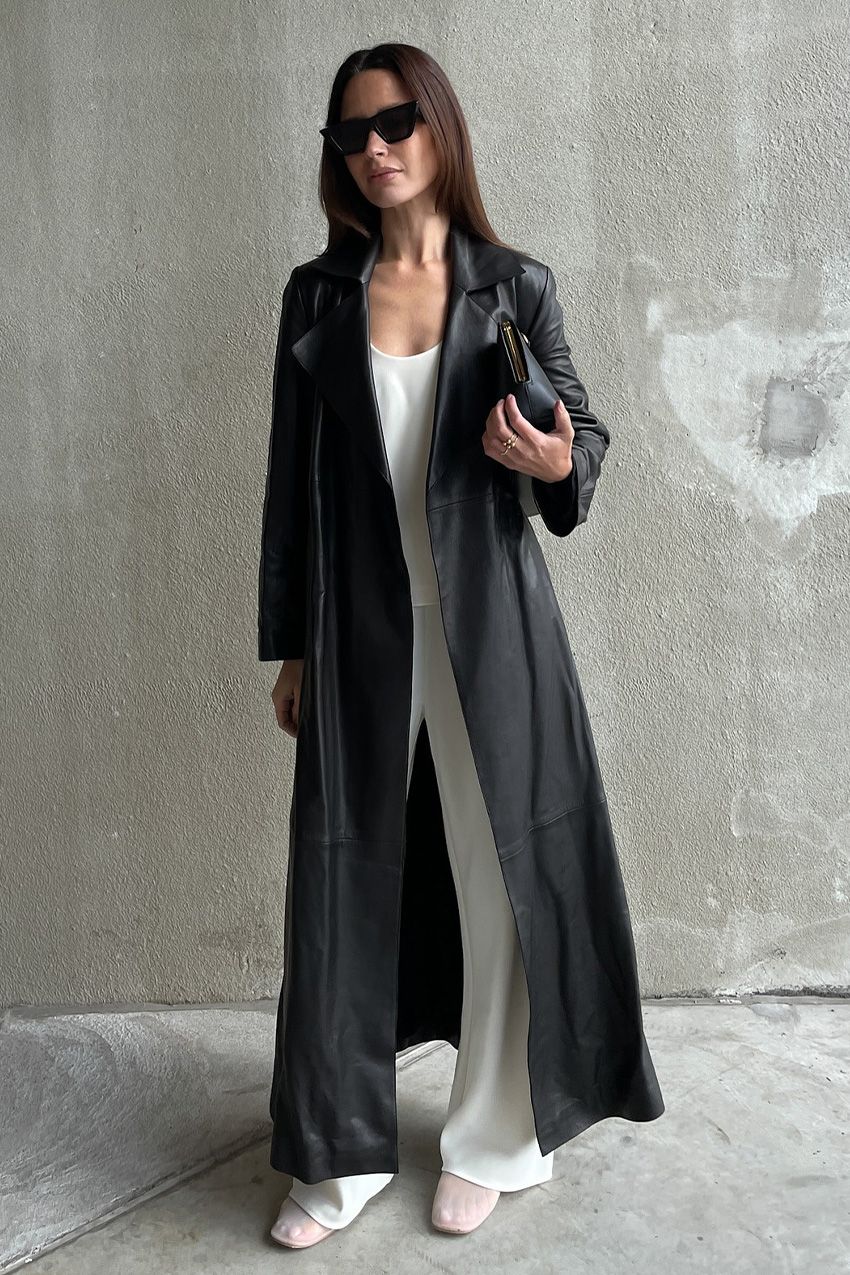 A woman standing outside in front of a concrete wall wearing a long leather trench on top of a white scoop neck tank, white wide-leg pants, white mesh ballet flats, a black clutch bag, and black cat-eye sunglasses.