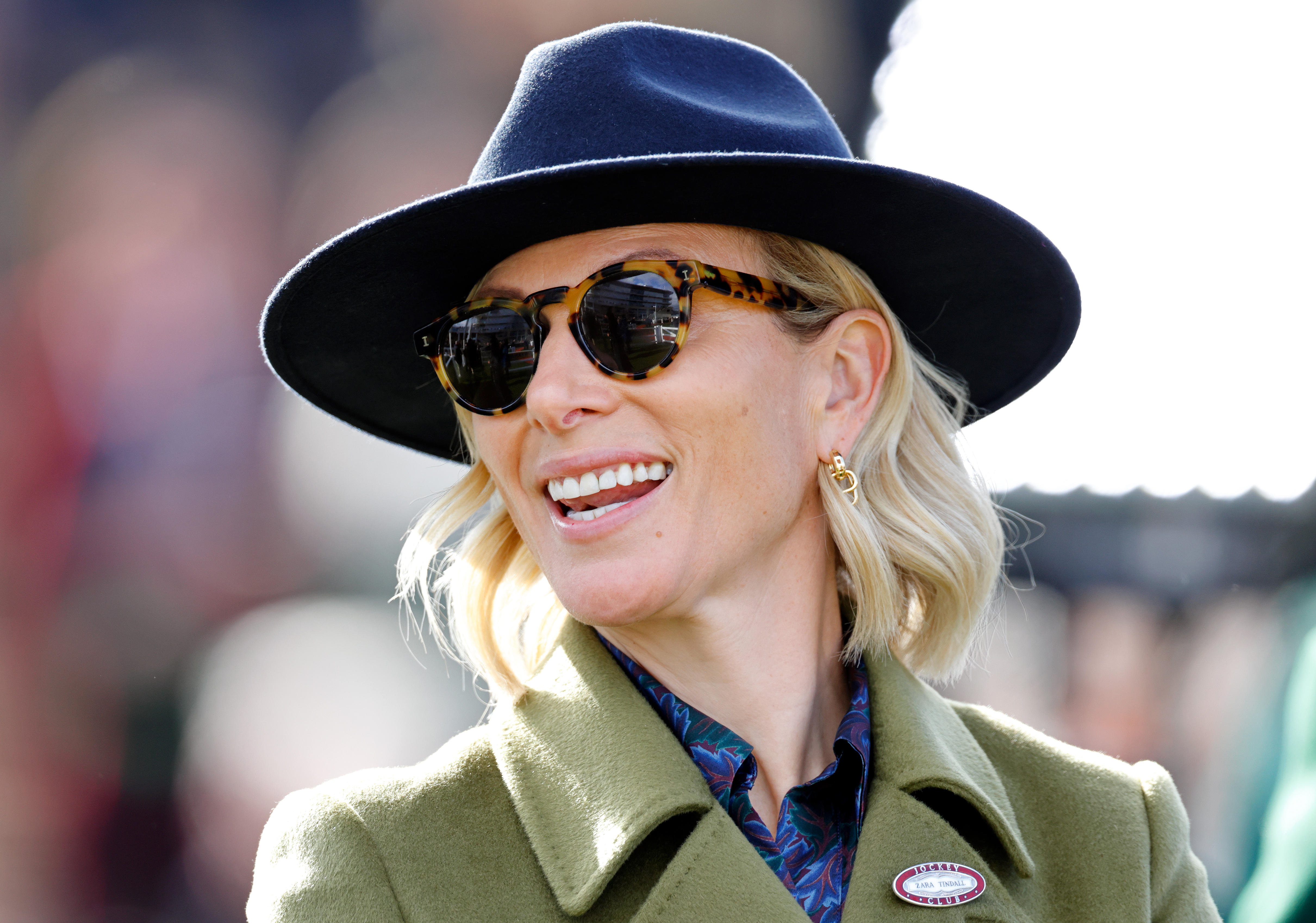 Zara Tindall wearing a blue fedora and sunglasses with a green coat and smiling