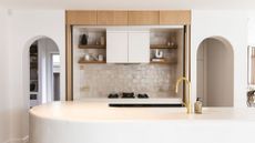 A large kitchen with doors, a long white island, brushed gold hardware, wooden shelving and a tile backsplash