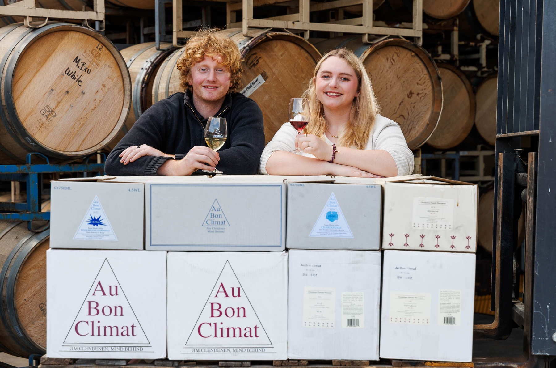 Siblings Knox Alexander and Isabelle Clendenen leaning on cases of Au Bon Climat wines in front of racks of barrels.