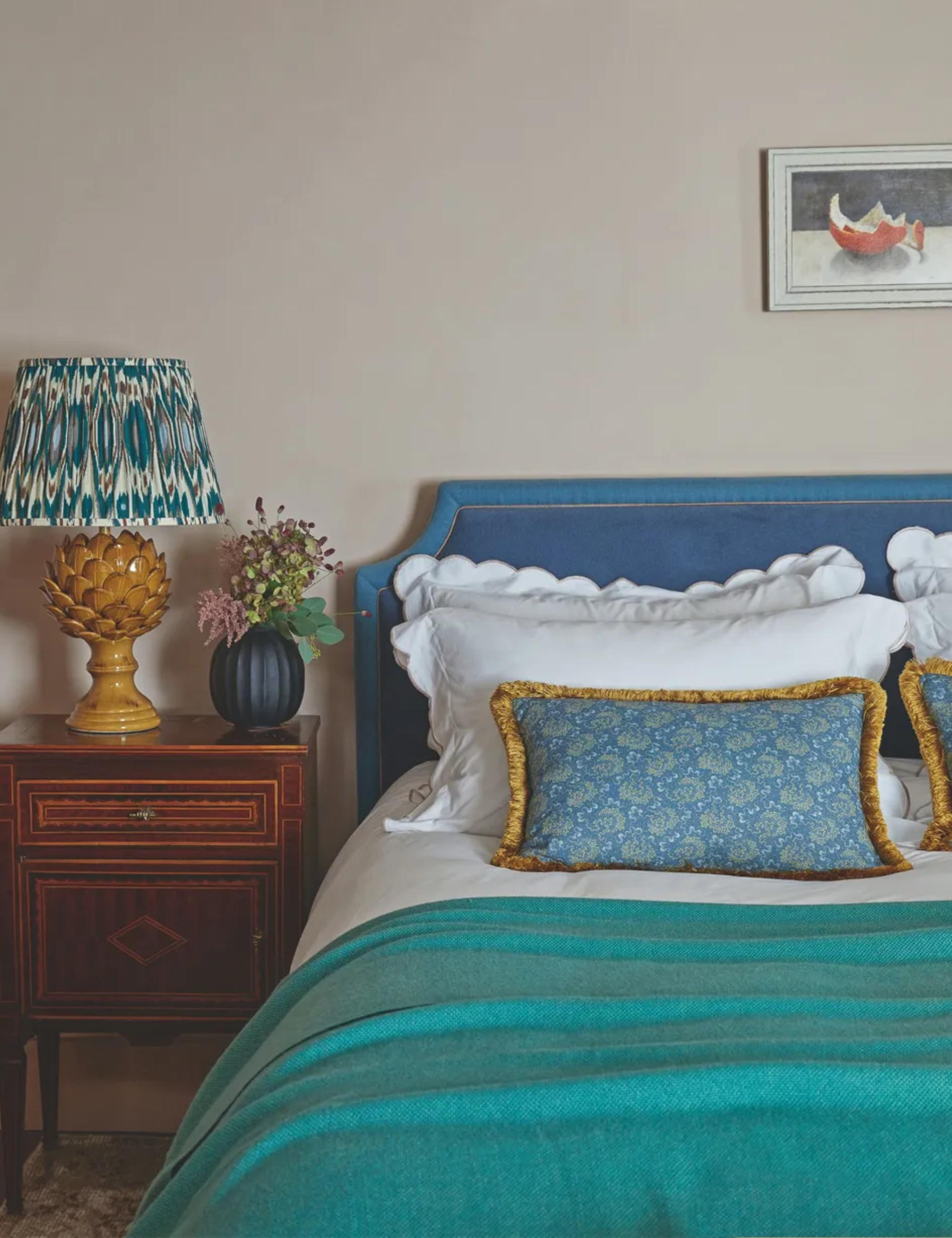A bedroom with a bright blue bed with blue bedding, a wooden bedside table, statement yellow ceramic lamp with patterned shade, small blue vase with flowers, neutral walls and a small still life artwork above the bed