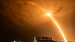 time lapse photo of an early morning rocket launch producing a bright streak arcing over a hangar and stretching beyond the clouds