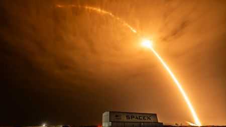 time lapse photo of an early morning rocket launch producing a bright streak arcing over a hangar and stretching beyond the clouds