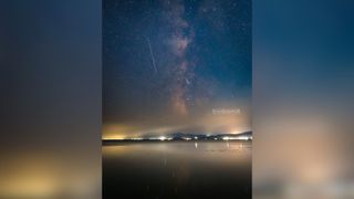 a vertical image shows a long perseid meteor streak in the sky with the milky way bulging in the center and a distant glow of yellow in the clouds.