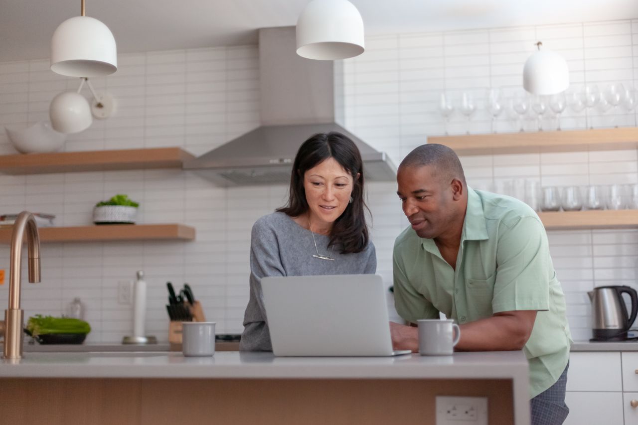 Couple stand at kitchen table as they look at laptop