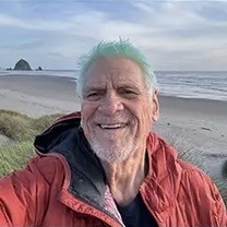 A middle aged man with teal hair is smiling in a red coat at the beach.