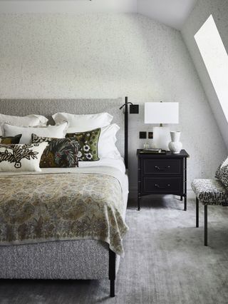 grey bedroom with grey carpet, grey textured bed, black bedside table, paisley throw, patterned cushions