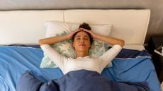 Woman lies on her back in bed, her head on a pillow. Here hands are on the top of her head and her eyes are closed. She looks calm and relaxed. 