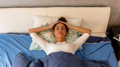 Woman lies on her back in bed, her head on a pillow. Here hands are on the top of her head and her eyes are closed. She looks calm and relaxed. 