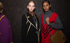 Two female models wearing looks from Prada's collection. One model is wearing a black and silver piece, brown neck scarf and black jacket with gold trims. Next to her is a model wearing a grey roll neck jumper with a red and black sleeveless piece over the top and she is holding a brown bag with chain handle. At the edge of the photo is a another model wearing a purple jacket with mustard coloured trims