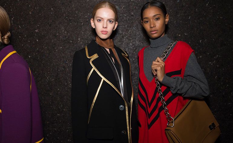 Two female models wearing looks from Prada&#039;s collection. One model is wearing a black and silver piece, brown neck scarf and black jacket with gold trims. Next to her is a model wearing a grey roll neck jumper with a red and black sleeveless piece over the top and she is holding a brown bag with chain handle. At the edge of the photo is a another model wearing a purple jacket with mustard coloured trims