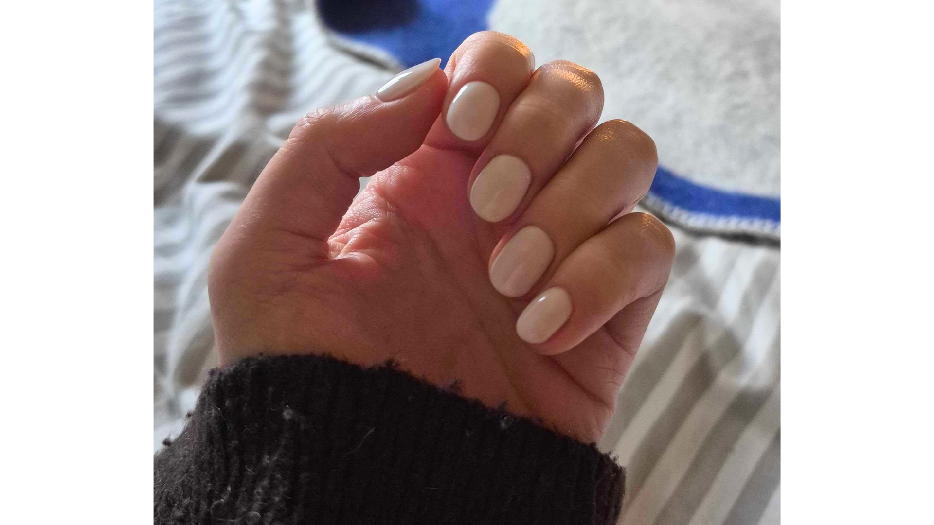 A close-up of Digital Beauty Writer, Naomi Jamieson's hand, which features short, milky white almond nails/ in front of a grey and white stripey bedding background