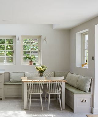 A white kitchen with a corner seating area with green seat cushions, gray and green throw pillows, a wooden table with two white chairs tucked underneath it, and three windows on the white walls