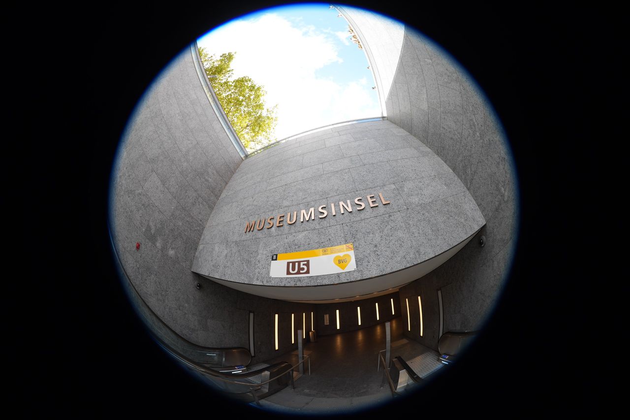 Fisheye view of the Museumsinsel U5 subway entrance in Berlin, with escalators descending beneath a curved stone wall.