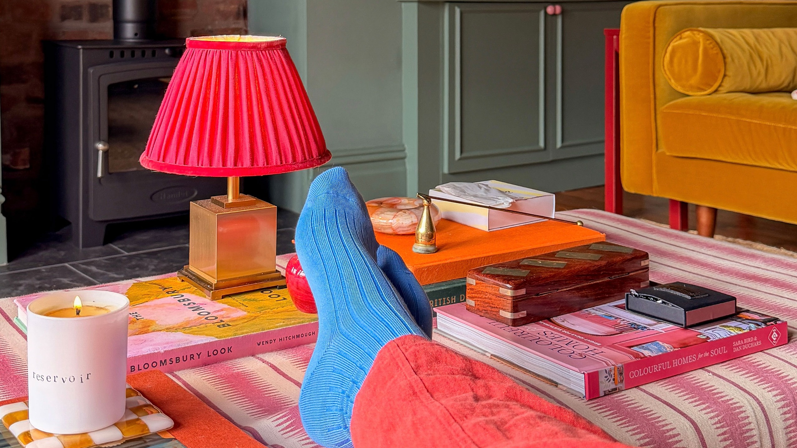 Woman in coral trousers and blue socks with feet resting on pink striped ottoman displaying candle, wireless lamp and stacks of coffee table books