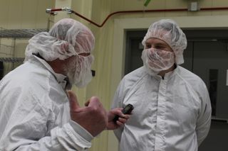 two men in white clean room suites face each other, likely conversing. the closer gentleman has his right thumb raised.