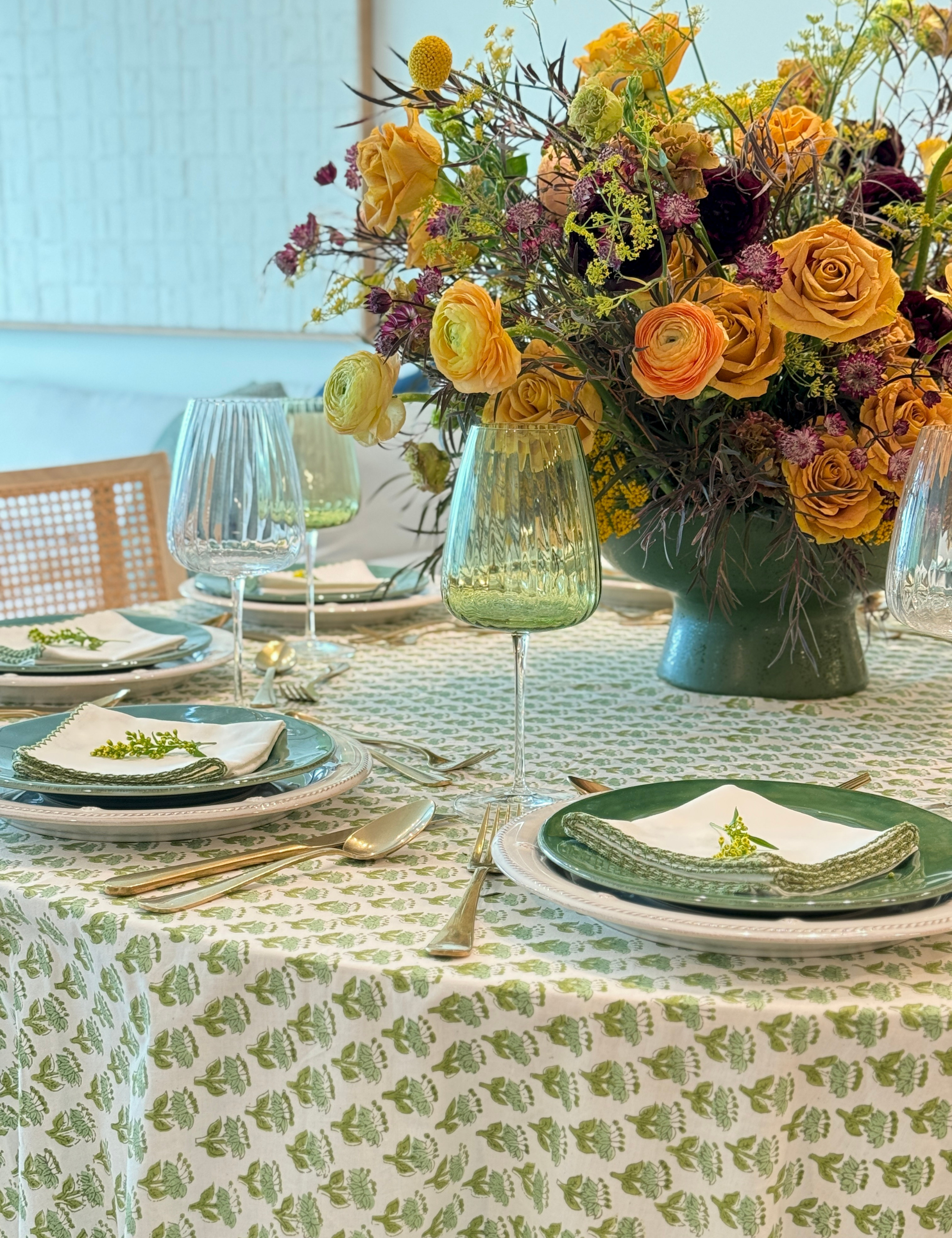 Fall tablescape featuring a green floral tablecloth, mix-and-match plate stacks, fluted wine glasses, and orange florals.