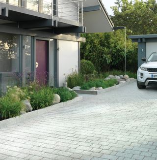 a grey stone driveway with planting by Stonemarket, next to a modern home with a white range rover to the right